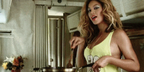 Beyoncé smiling in her "Party" music video holding pan with one hand and a wooden spatula with the other making stirring moves